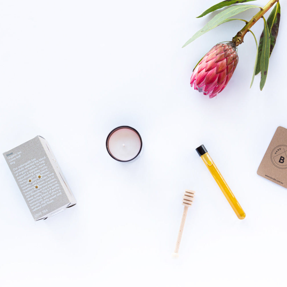 Assorted items including a notebook, candle, pencil, and card on a white surface with a pink flower for Bindle gift hamper